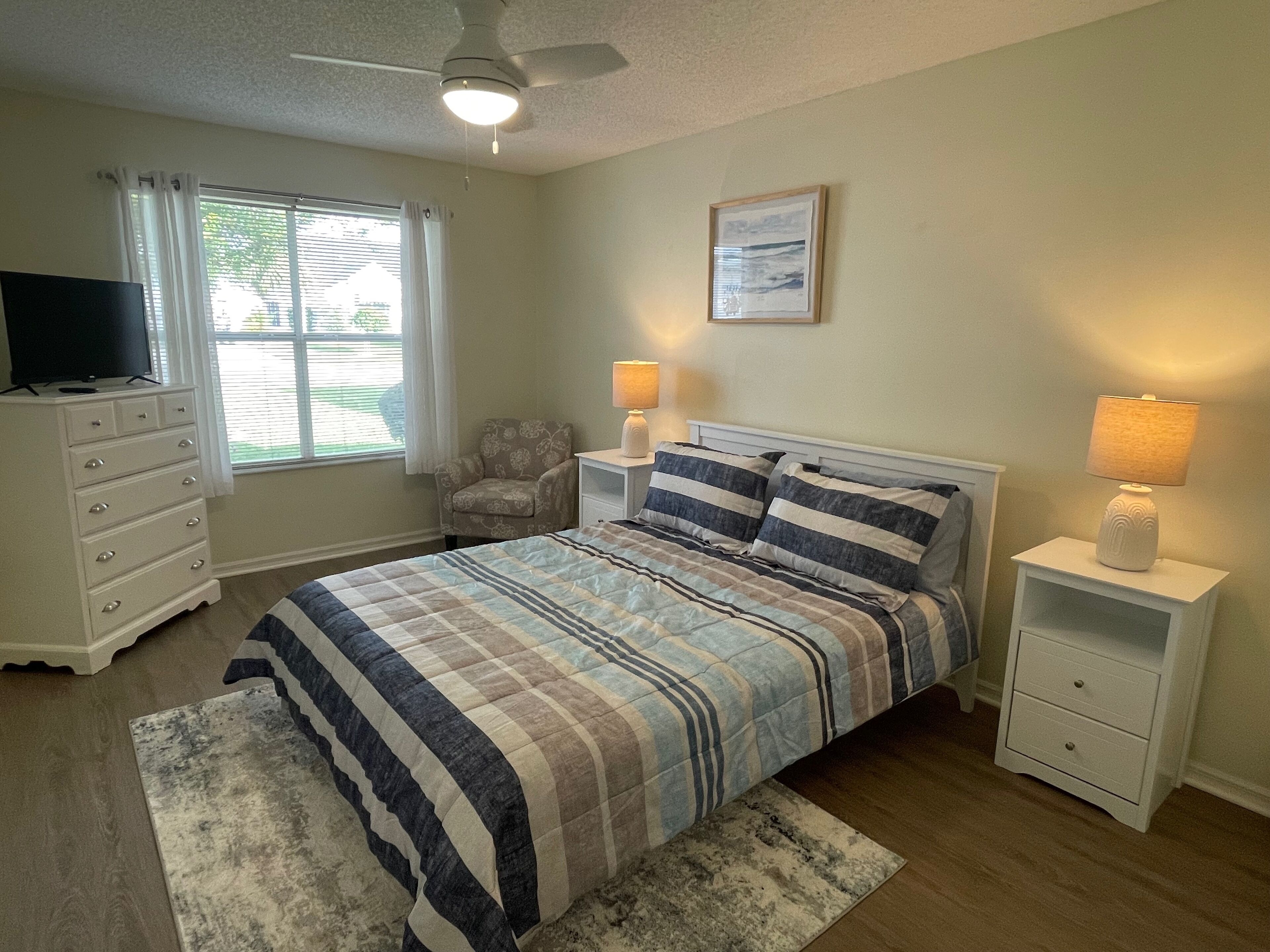 Une chambre à coucher comprenant un lit, une commode, une chaise, une table de chevet avec une lampe et un ventilateur de plafond.