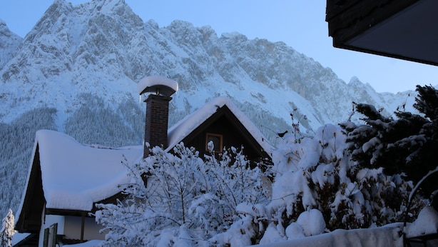 A mountain cabin with a snow-covered roof and chimney, surrounded by snow-covered trees and mountains.
