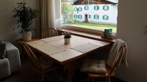 A dining area with a wooden table, chairs, a potted plant, and a view of a mountain house through a window.