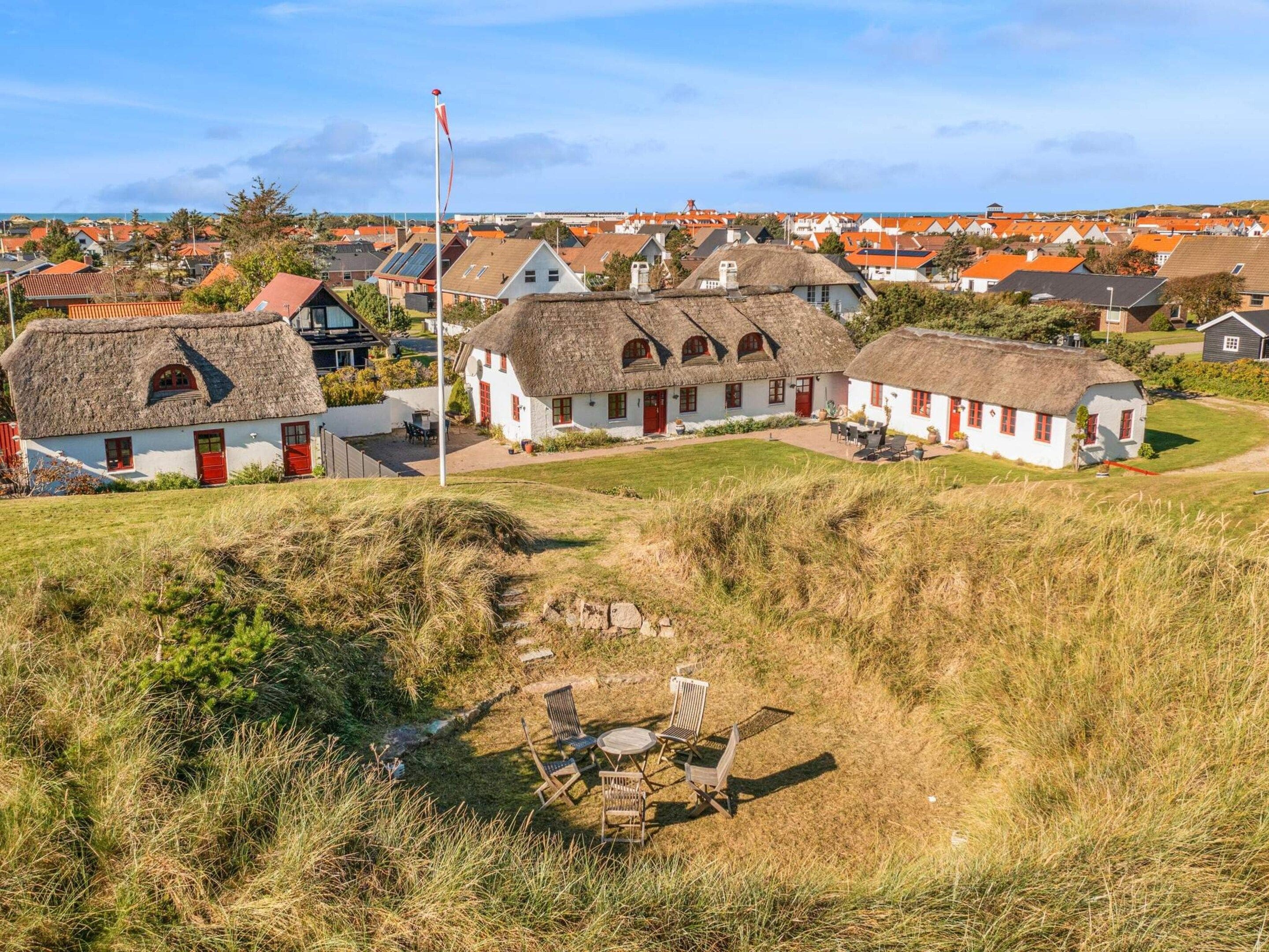 Un pueblo rural con casas tradicionales de techo de paja, un asta de bandera y un espacio verde con muebles de exterior.