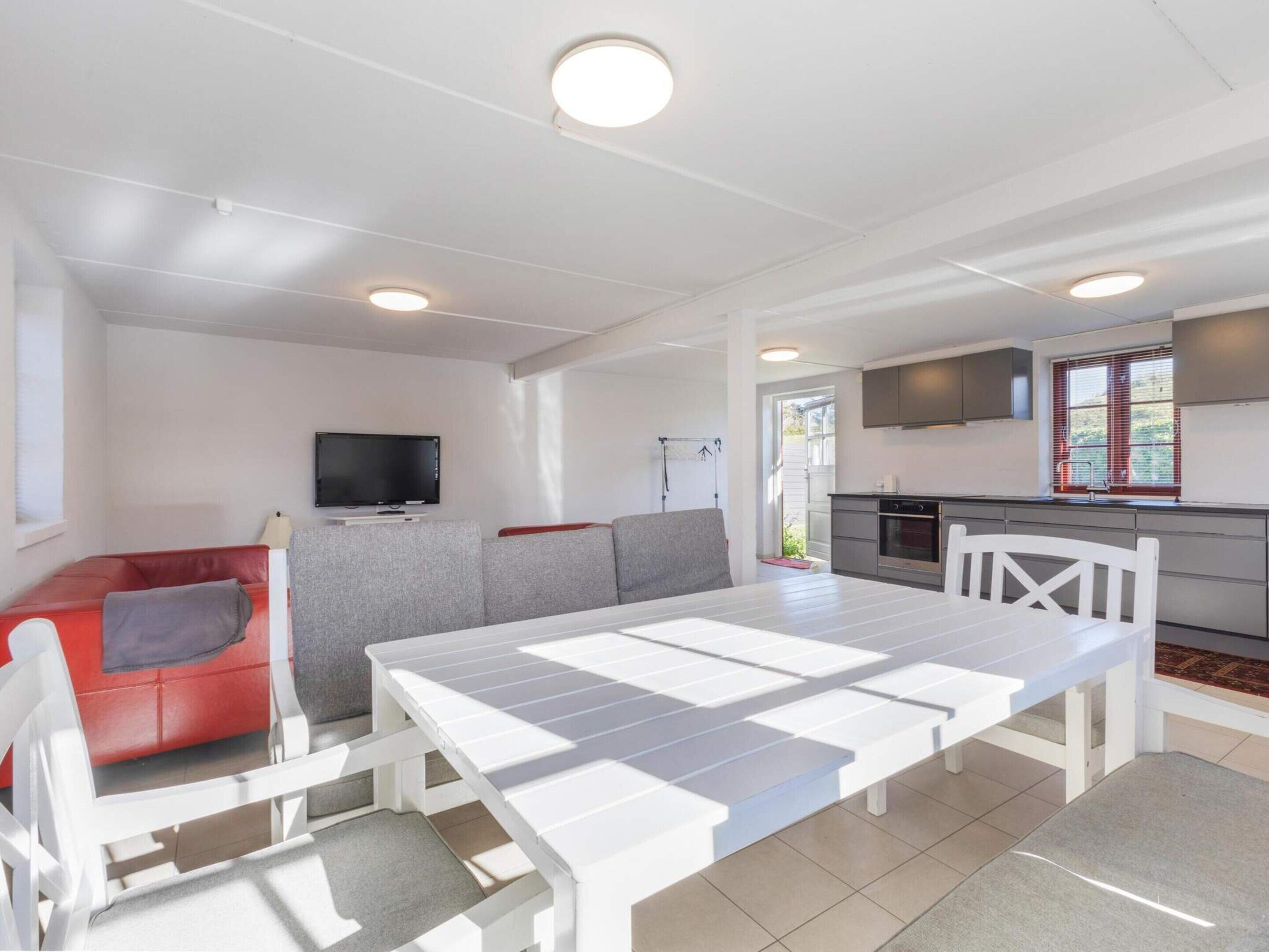 A modern living space with a white dining table, red sofa, grey sectional, and a kitchen area with a TV and window.