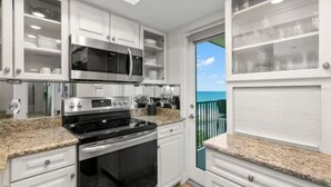 A modern kitchen with white cabinetry, stainless steel appliances, and a view of the ocean through a sliding glass door.