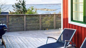 A wooden deck with a view of the sea, a barbecue grill, and a red building with a window. - 4 Star Holiday Home in Branno (Branno)