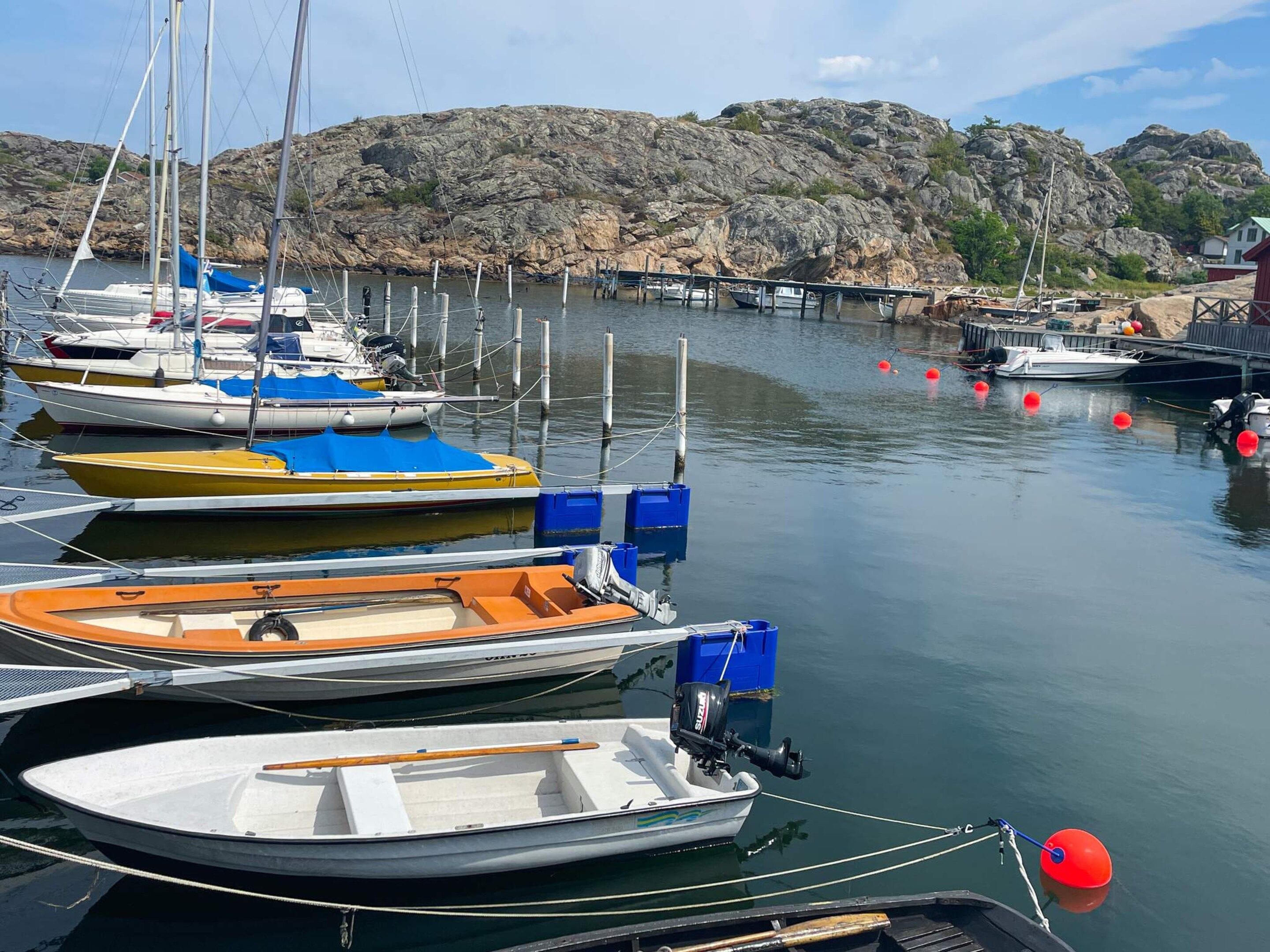 Uma marina com vários barcos atracados, uma paisagem rochosa ao fundo e um pequeno edifício à direita.