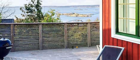 Una terraza de madera con vistas al mar, una parrilla y un edificio rojo con una ventana.