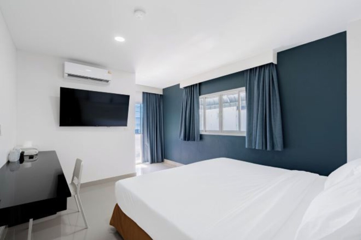 A modern hotel room with a bed, a flat-screen TV mounted on the wall, a desk with a chair, and a window with curtains.
