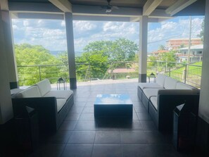 A spacious balcony with two white sofas and a central coffee table, offering a view of a green landscape and buildings in the distance. - Casa Ellysian (San Ignacio)