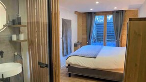 A bedroom with a bed, wooden flooring, a window with curtains, and a bathroom with a sink and mirror. - Cosy chalet at the foot of the bossons glacier opposite the ski shuttles (Chamonix-Mont-Blanc)