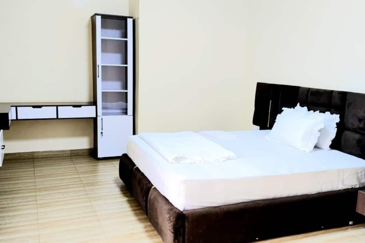 A neatly made bed with a black headboard, a bedside table with a drawer, and a white wardrobe with open doors.