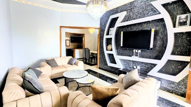 A modern living room with a beige sofa set, a round coffee table, and a large TV mounted on a black and white geometric wall. - Silver Budget & Travel Accommodation (Kigali)