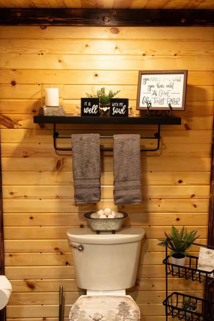 Ein Badezimmer aus Holz mit Toilette, zwei Handtüchern an einem Halter, einer Ablage mit Kerze und einer kleinen Pflanze sowie einem Schild mit einer Botschaft.