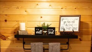Une salle de bain en bois avec des toilettes, deux serviettes suspendues à un porte-serviettes, une étagère avec une bougie, une petite plante et un panneau avec un message.