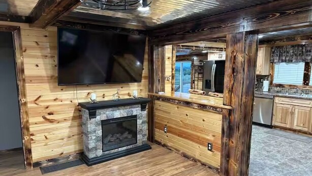 Un intérieur rustique avec une cheminée en pierre, des murs en bois, un téléviseur fixé au mur et un coin cuisine visible à travers une porte ouverte.
