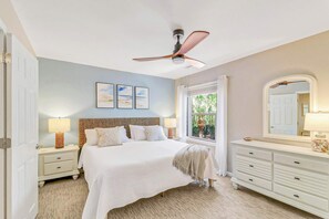 Quarto com cama, mesas de cabeceira, penteadeira, ventoinha no teto e quadros nas paredes.
