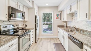 Una cocina moderna con armarios blancos, electrodomésticos de acero inoxidable, encimera de granito y una ventana con persianas.
