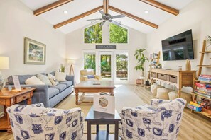 A spacious living room with a high ceiling, a ceiling fan, a large TV, and a view of the outdoors through glass doors.