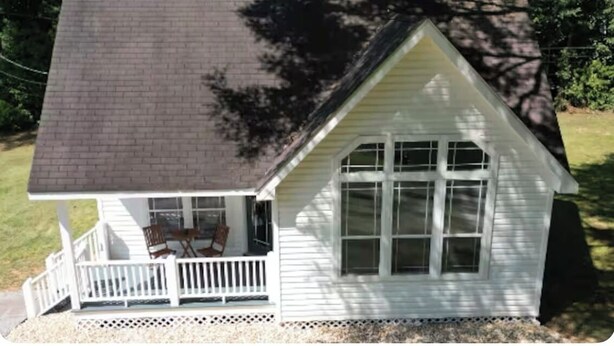 A small white house with a dark roof, a porch with chairs, and a large window.