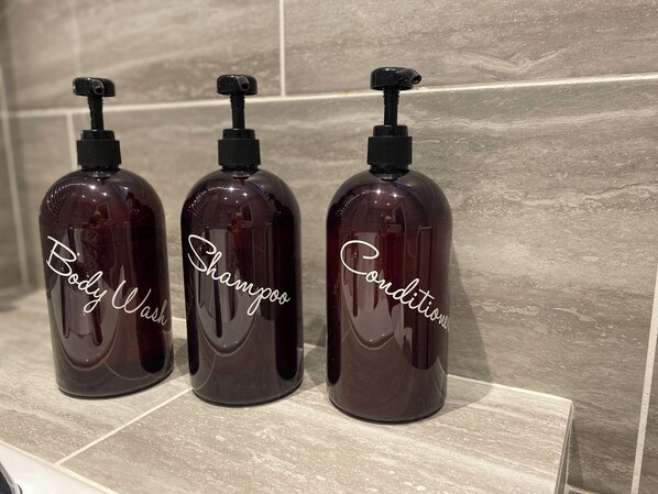 Three dark brown bottles with pumps labeled "Body Wash," "Shampoo," and "Conditioner" on a tiled surface.