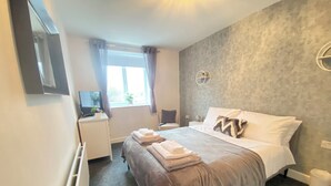 A modern hotel room with a bed, bedside tables, a desk, a window with curtains, and a wall-mounted light fixture.