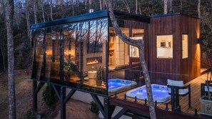 A glass-enclosed cabin with a hot tub and a wooden structure adjacent to it, surrounded by trees.