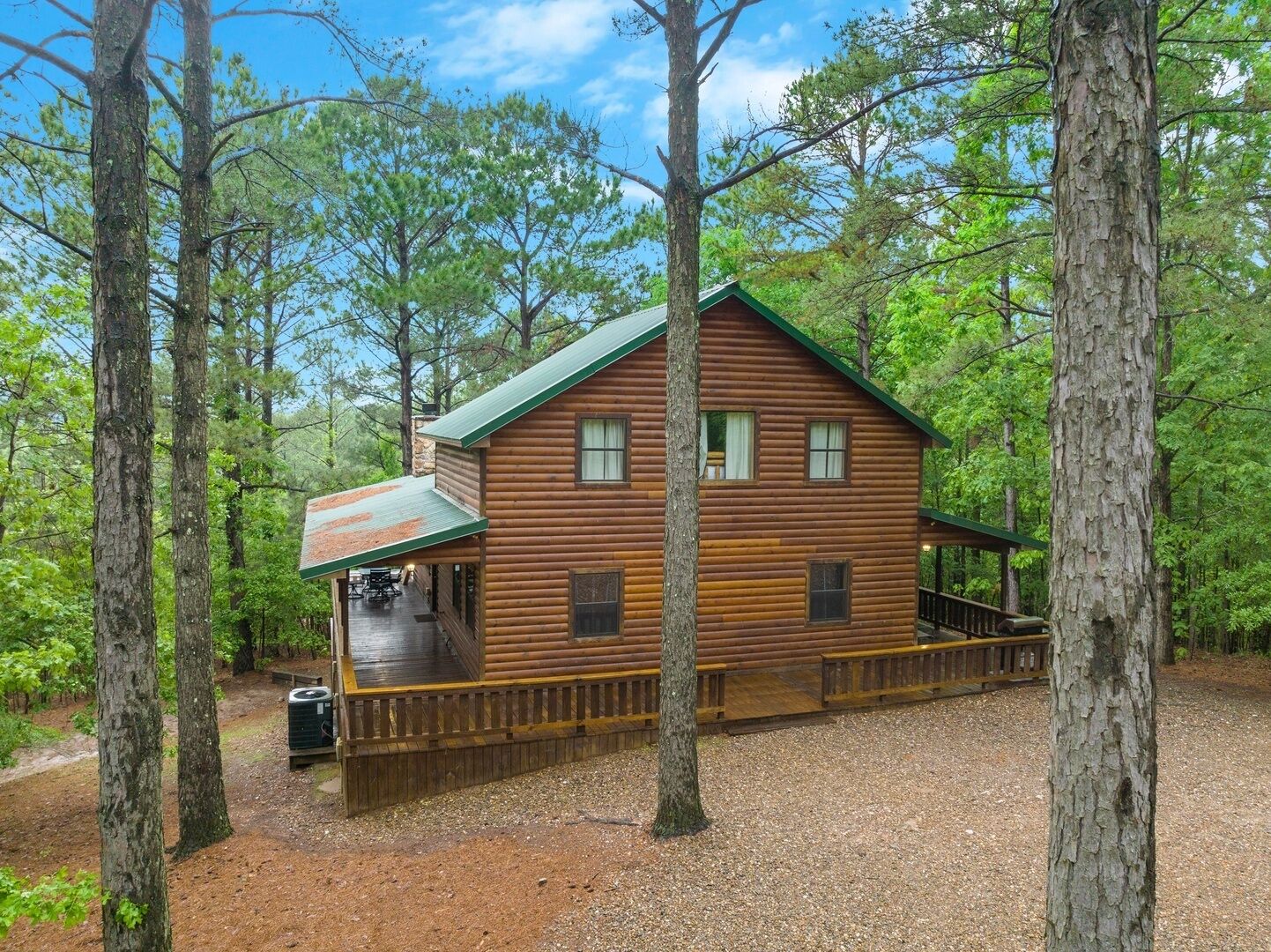 Une cabane en rondins dotée d’un porche couvert et d’une terrasse clôturée, entourée de grands arbres.
