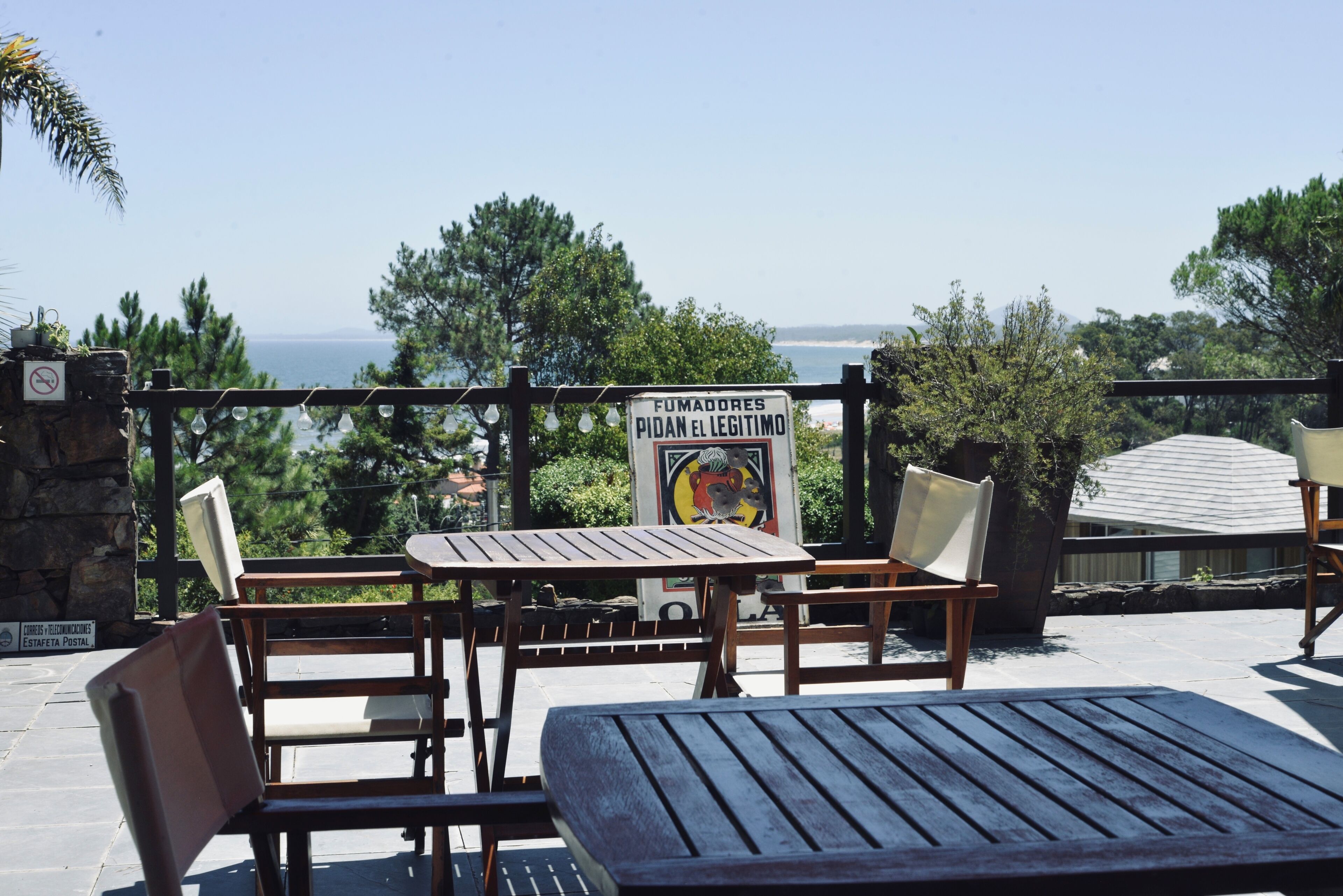 An outdoor seating area with wooden tables and chairs, a sign with a warning, and a view of the sea.