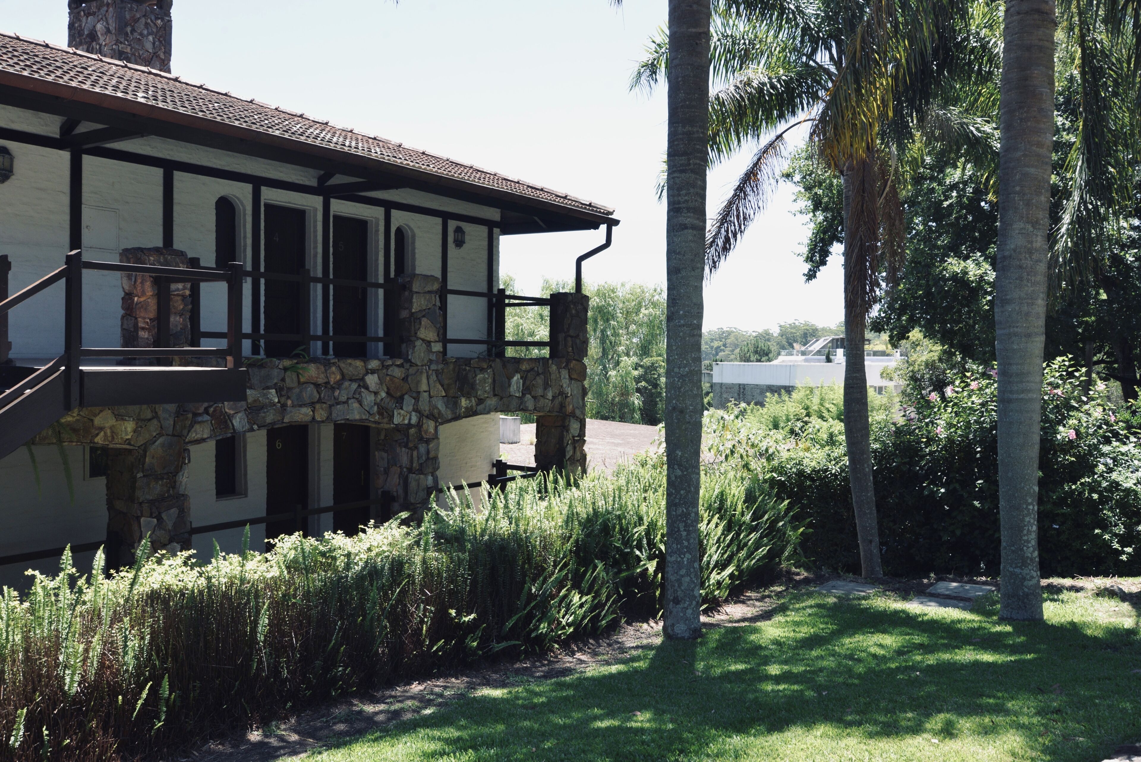 A two-story stone house with a balcony, surrounded by lush greenery and palm trees.