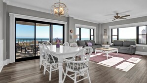 A spacious living room with a dining area, large windows offering ocean views, and a ceiling fan. - Wrightsville Oceanfront Condo: Pool, Ocean Views, Beach Access (Wrightsville Beach)