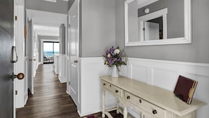 A hallway with a wooden floor, white wainscoting, a console table with a vase of flowers, and a mirror on the wall. - Wrightsville Oceanfront Condo: Pool, Ocean Views, Beach Access (Wrightsville Beach)