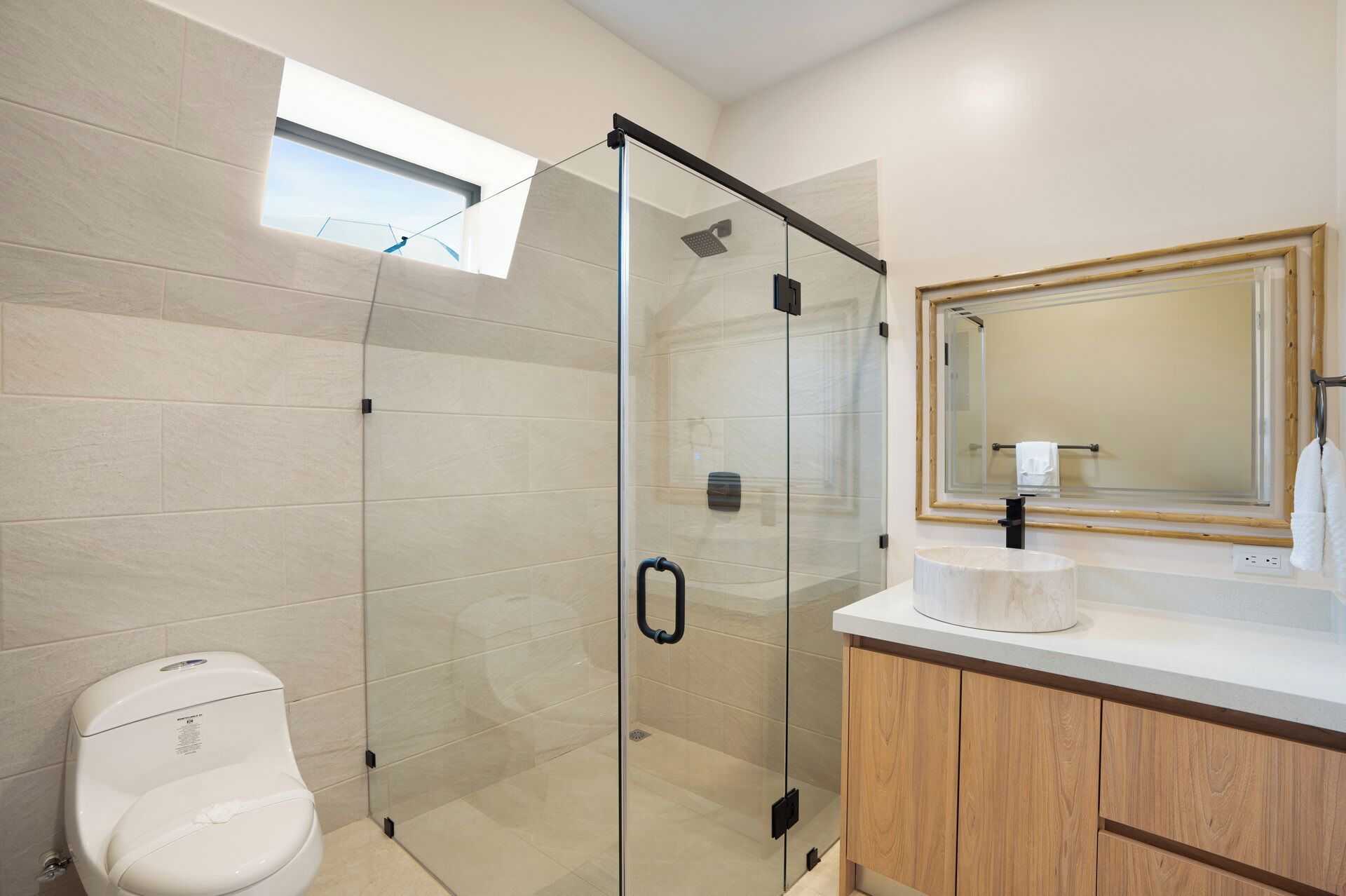 A modern bathroom with a glass-enclosed shower, a toilet, a wooden vanity with a mirror, and a window above the shower.