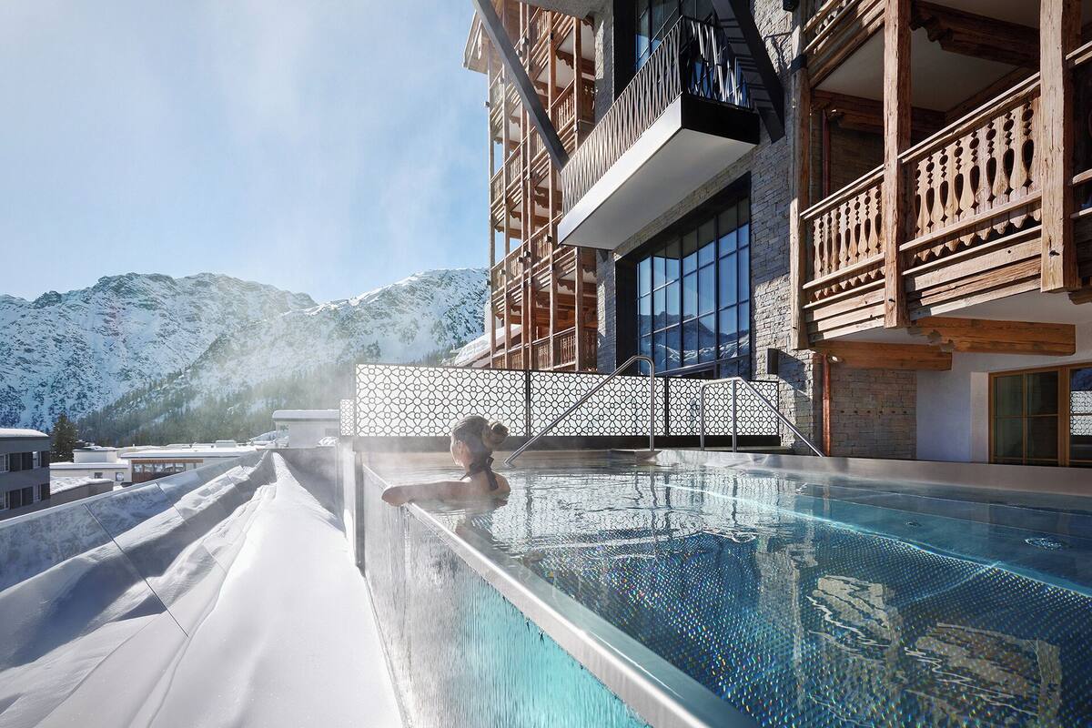 A person in a pool with a mountain backdrop, wooden balconies, and a modern building facade.