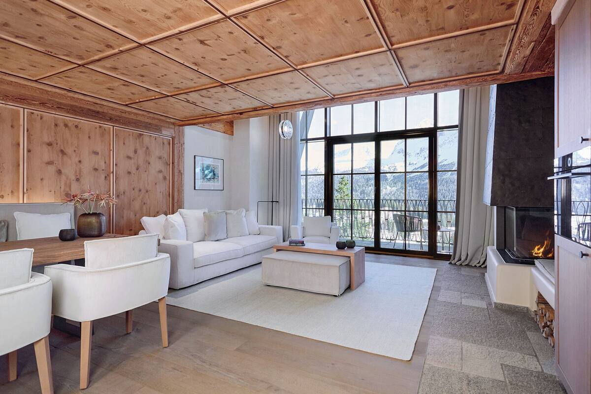A modern living room with a wooden ceiling, white furniture, a fireplace, and a large window with a mountain view.