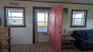 Una habitación con suelo de madera, una puerta roja, un sofá azul y dos ventanas con vistas al mar.
