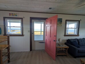 A room with a wooden floor, a red door, a blue sofa, and two windows with views of the sea.