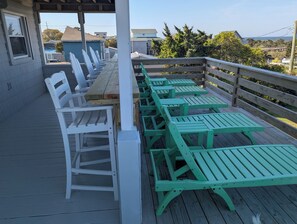 A covered outdoor seating area with wooden tables and chairs, a wooden railing, and a view of the surrounding neighborhood.