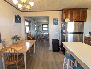 A kitchen with wooden cabinets, a stainless steel refrigerator, a dining table with chairs, and a trash can.