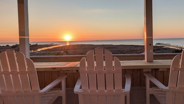 Tres sillas Adirondack blancas mirando hacia una puesta de sol sobre el océano, con una terraza de madera y un muelle al fondo.