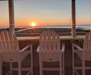 Tres sillas Adirondack blancas mirando hacia una puesta de sol sobre el océano, con una terraza de madera y un muelle al fondo.