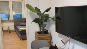 Ein modernes Wohnzimmer mit einem grauen Sofa, einem runden Tisch mit Lampe, einer großen Topfpflanze und einem flachbildfernseher an der Wand.