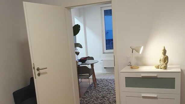 Ein Schlafzimmer mit einem Bett, einem Nachttisch mit Lampe und Blick in einen weiteren Raum mit Stuhl und Pflanze.