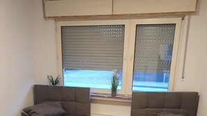 Ein Zimmer mit zwei Betten, einem Fenster mit Jalousien, einem Heizkörper und einer kleinen Pflanze im Fensterbrett.