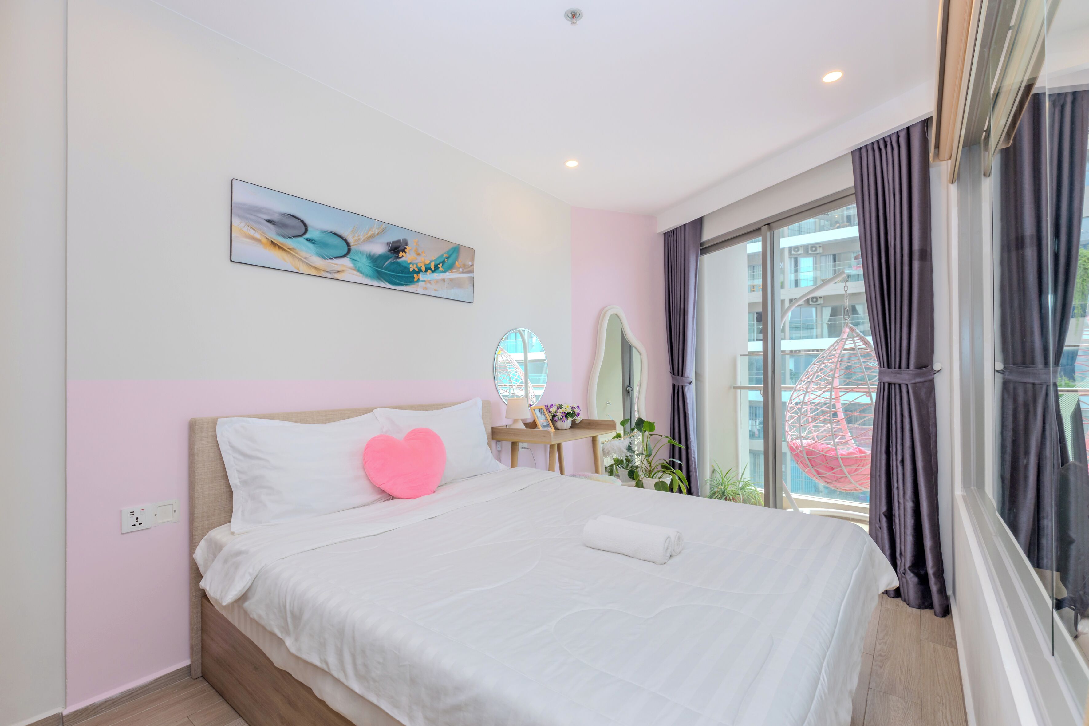 Un lit bien fait, avec une literie blanche et un coussin en forme de cœur rose, un tableau abstrait accroché au mur, une petite table avec une lampe et des fleurs, et une grande fenêtre avec des rideaux.