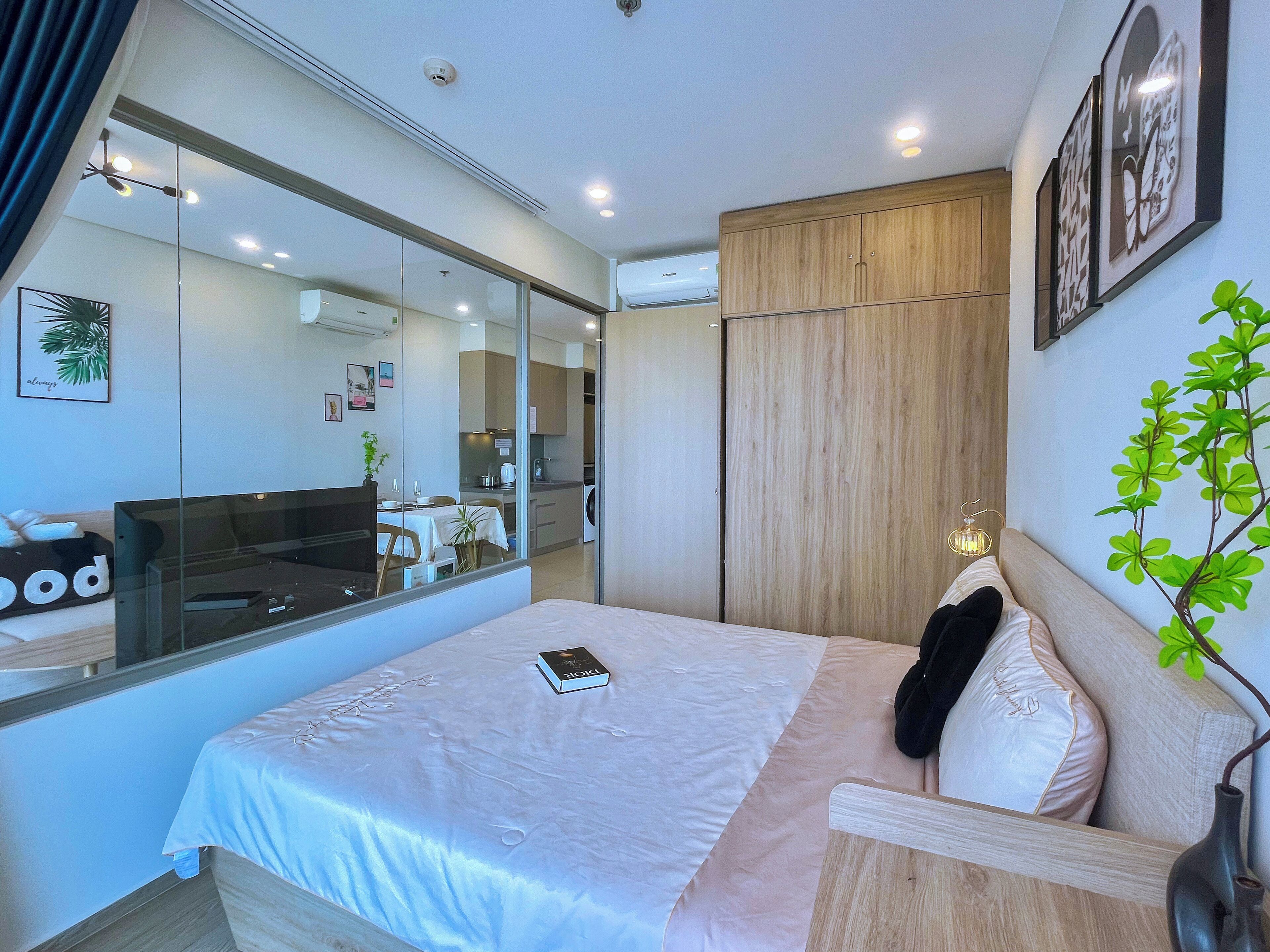 A modern hotel room with a large bed, a bedside table, a mirror, a wardrobe, and a view of the kitchen and living area.