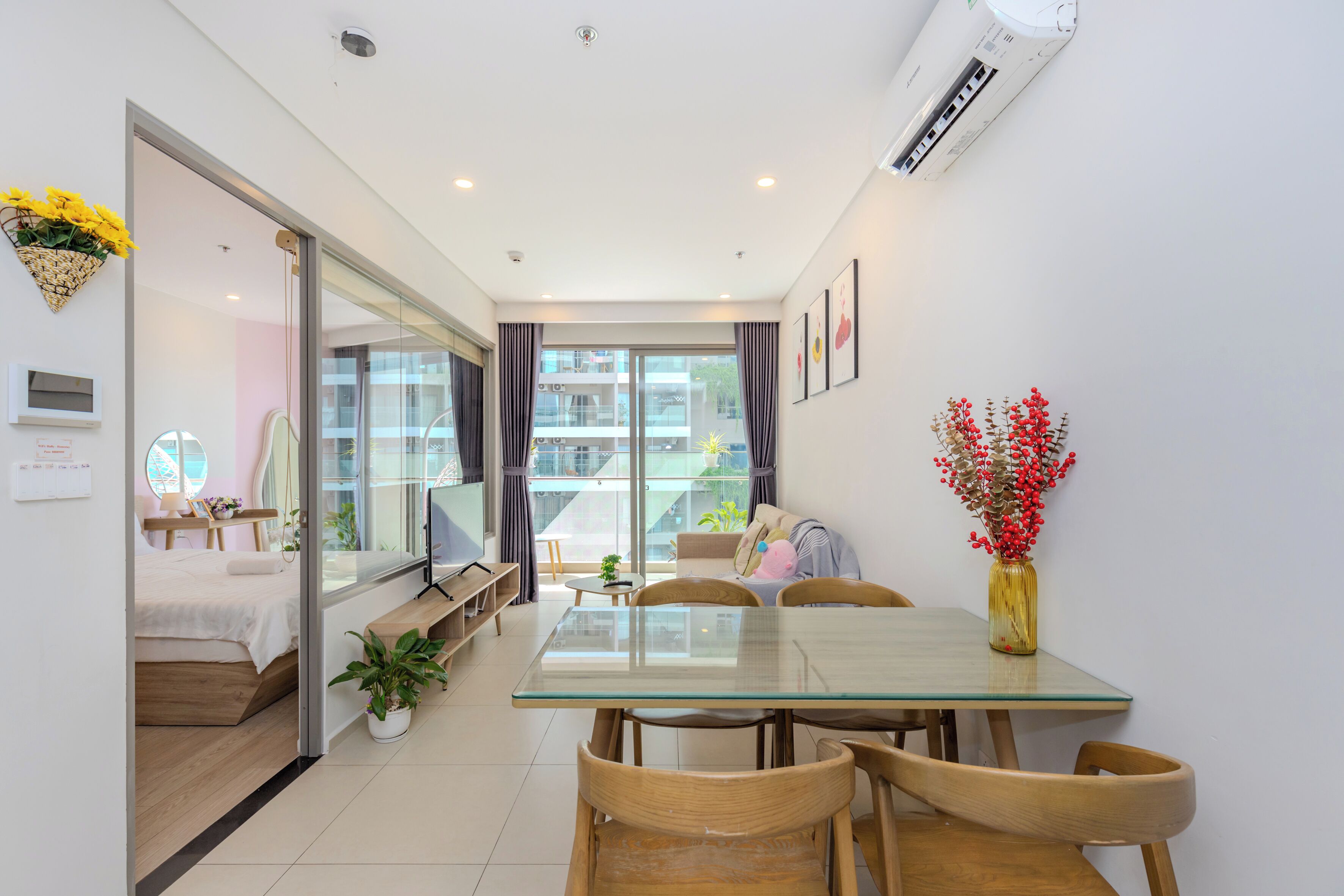 A modern apartment with a glass dining table, wooden chairs, a sofa, a bed, and a balcony with a view.