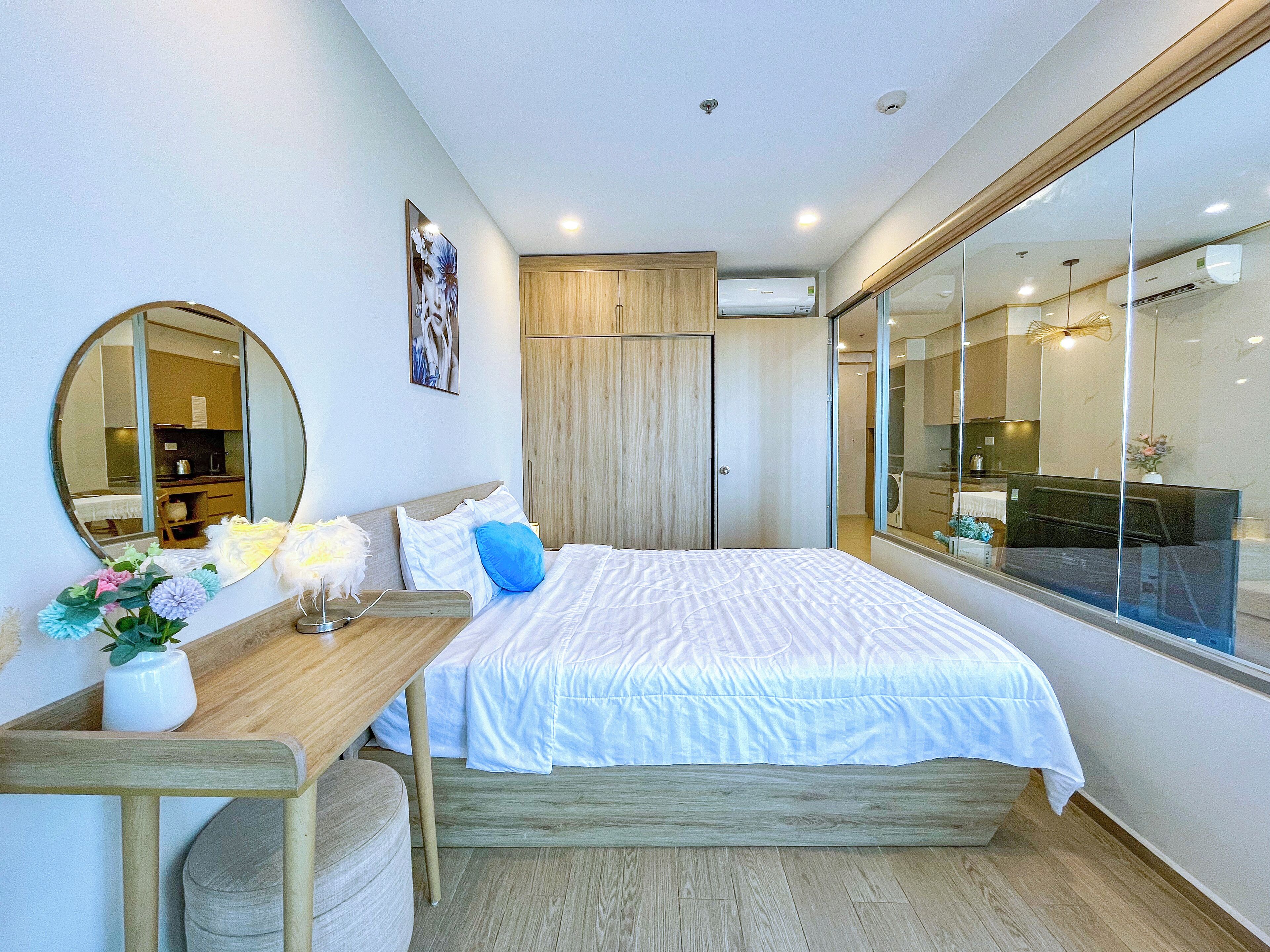 A modern bedroom with a wooden bed, a desk with a lamp, a mirror, and a view of the kitchen through a glass door.