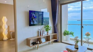 A modern living room with a large flat-screen TV mounted on the wall, a wooden console table with books and decorative items, a small round table with a vase, and a view of the sea through a large glass door. - OLIU'S HOMESTAY - The Sóng Vũng Tàu (Ho Chi Minh City)