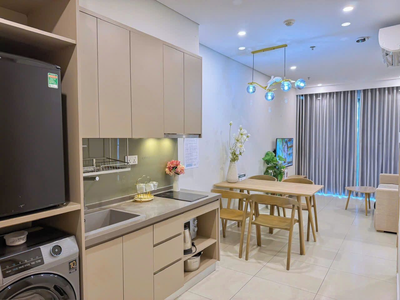 Een moderne keuken met een ingebouwde wasmachine, een eethoek met houten meubels, en een woonruimte met een televisie en gordijnen.