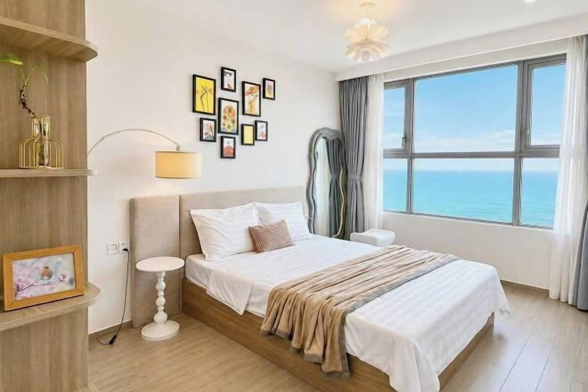 Une chambre d’hôtel avec un lit, des tables de chevet, une lampe, un mur orné de tableaux, une grande fenêtre donnant sur un beau paysage et une étagère en bois.