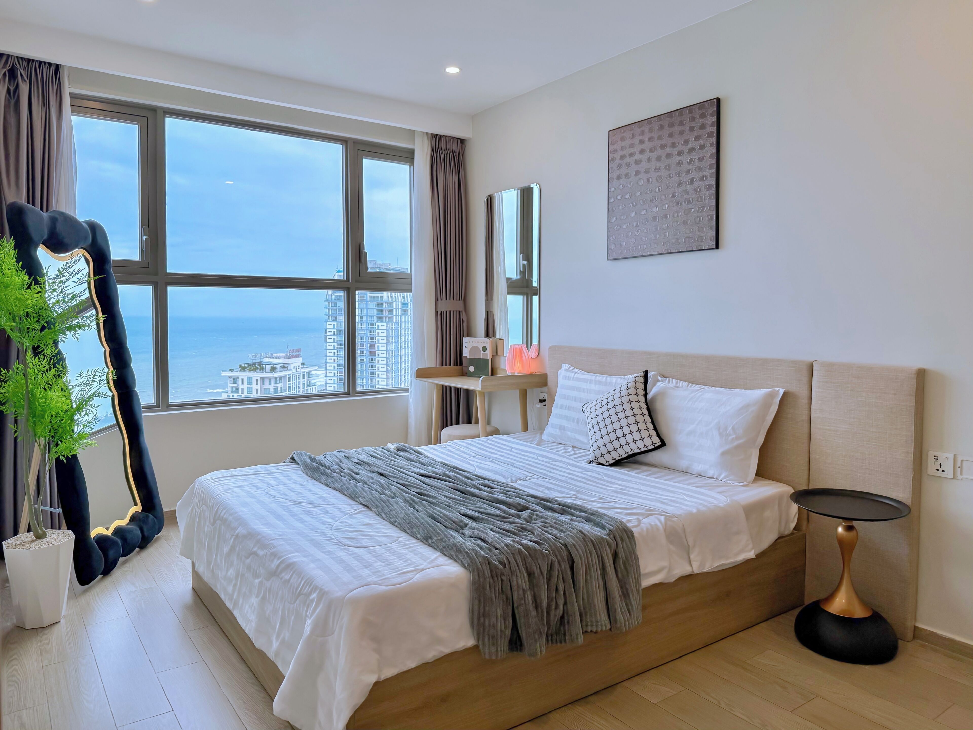 A hotel room with a large window, a bed with white bedding, a bedside table, a mirror, and a potted plant.