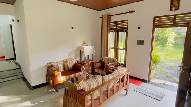 A spacious living room with a wooden ceiling and a ceiling fan, a sofa set, a small table with a lamp, and a view of greenery outside. - Riverside Nature Haven Villa  (weligama)